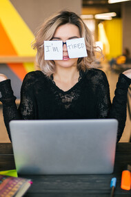 A portrait of a young woman sitting at a table, paper stickers stuck to her glasses, her expression tired and worn