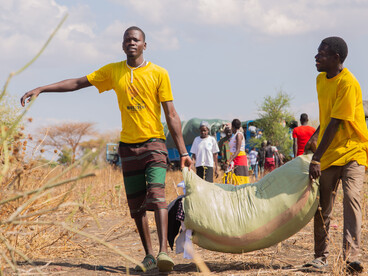 A man carries humanitarian aid across a rural landscape in South Sudan, with a truck and local residents in the background
