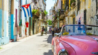A vintage pink convertible taxi rolls through a colorful Havana street under the Cuban flag, evoking a vivid portrait of resilience, history, and the everyday life shaped by decades of geopolitical tension and economic isolation