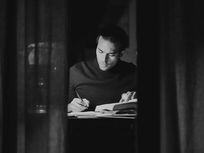 Man in black sweater in black and white room deep in thought and writing book