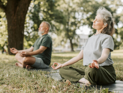A gray-haired man and a woman meditating, sitting on mats, in a park