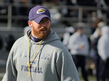 An American football coach in a blue and white jacket and black helmet shouts directions from the sidelines
