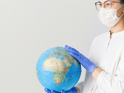 Mulher segura globo terrestre com máscara e luvas de proteção. A questão que muitos especialistas tentam responder é: os sintomas de longo prazo são causados pelo próprio vírus, pela vacina ou por uma interação entre os dois? Ainda não há uma resposta definitiva, mas estudos continuam sendo conduzidos