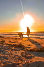 A man walks his dog along the beach as the sun sets over the horizon