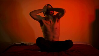A topless man sits alone on his bed, his posture reflecting anguish and anxiety in a quiet, dimly lit bedroom