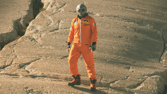 An alien humanoid figure in an orange jumpsuit looks at the camera