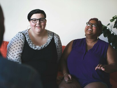 Two plus-size women are sitting comfortably on the couch, sharing a conversation and smiling at each other