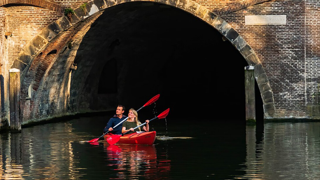 A man and woman paddle a red kayak along a river in Utrecht, the Netherlands, under the bright daytime sky