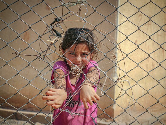 A young girl grips a chain-link fence in Idlib, Idlib Governorate, Syria