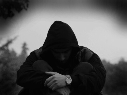 A man crouching on the ground wearing a hoodie trying to conceal the absence of his happiness