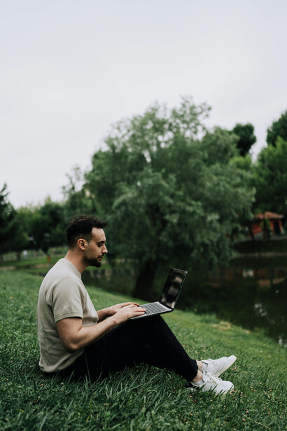 Sitting on the grass near a scenic pond in a park, a man works on his laptop, blending productivity with a calm outdoor atmosphere