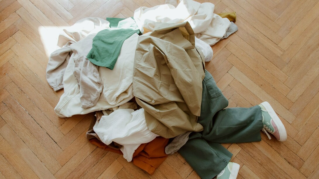 Diversas peças de roupa cobrem completamente uma pessoa deitada no chão. A imagem simboliza o excesso de consumo e convida à reflexão sobre a quantidade de vestuário adquirida e descartada atualmente. O cenário representa o impacto da cultura do consumo acelerado e a necessidade de escolhas mais conscientes