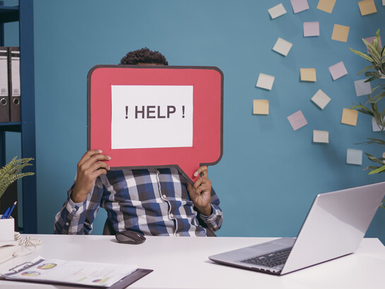 A worker raising a “HELP!” message while seated at a computer, symbolizing how a need for assistance can become an opening for social engineering