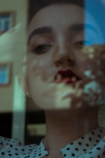 A woman in a blue and white polka dot shirt gazes out from behind a window, caught between emotion and reality