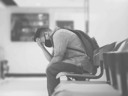 A man overwhelmed with the amount on his to-do list is sitting in a public room wearing a protective mask