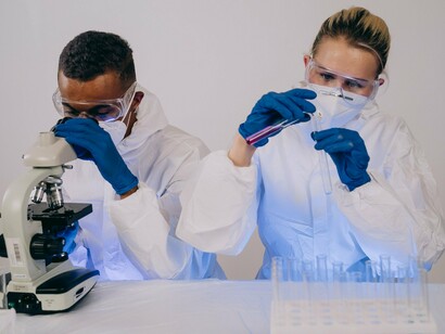 Dois pesquisadores com equipamentos de proteção trabalham com um microscópio e tubos de ensaio em um laboratório. A investigação de novas terapias contra o melanoma envolve o estudo de características moleculares dos tumores e de estratégias para estimular o sistema imunológico a reconhecer as células cancerosas
