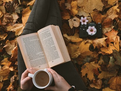 A person enjoying a cup of coffee while reading a book, seated among autumn leaves