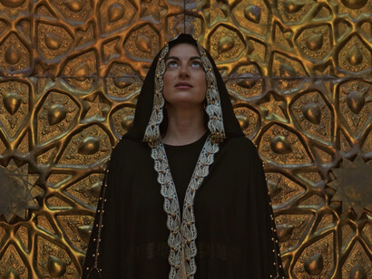 Standing before the golden doors of the mosque, she symbolises the hope and transformation sought by women in contemporary society, echoing Mosteghanemi's narratives of love and autonomy