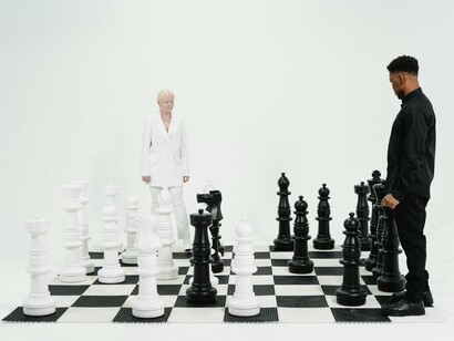 A man and a woman stand in a tense standoff on a giant chessboard, blending seamlessly with the black and white pieces, highlighting the calculated tension of strategy and rivalry. 