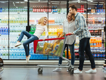 Smiling parents and their child sit on a cart in a brightly lit supermarket aisle, surrounded by colorful food displays
