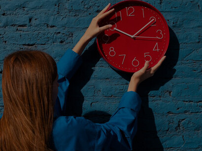 A woman holding a wall clock amidst a backdrop of intricate complexity diagrams and clock faces, showcasing the interplay of timepieces and the complexity of time itself