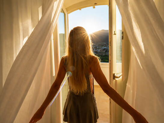 Golden light spills through the open doorway as a girl welcomes the morning, caught between quiet solitude and the promise of a new day