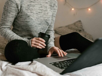 A woman in a gray sweater sits on a bed with books and a laptop in a minimalist room