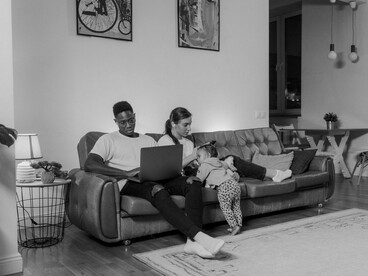 Sentados ao lado da filha, os pais utilizam computador e celular simultaneamente. A crescente influência da tecnologia é apresentada como um dos elementos que transformaram as dinâmicas familiares contemporâneas