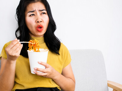 A woman struggles with the overwhelming heat of extra-spicy noodles, her flushed face and burning tongue capturing the powerful sensation of the meal