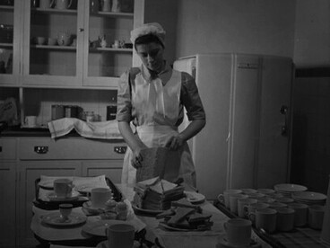 Estudiante de enfermería corta pan para sus pacientes en la cocina de la sala del Hospital St Helier. Está preparando té, huevos duros y pan con mantequilla. 1943