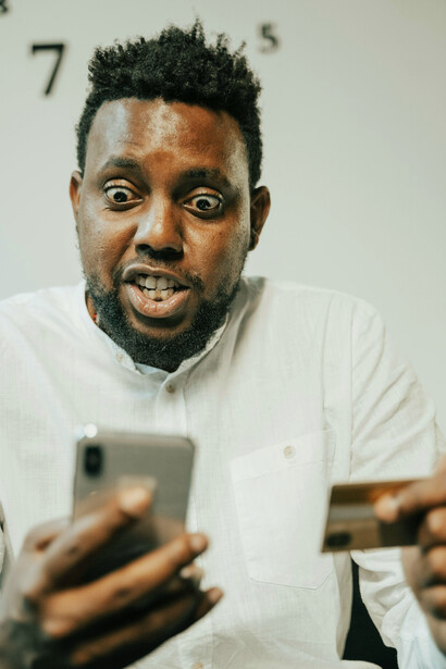 A man in a white long-sleeved shirt holding a phone and a card, reacting to shocking email news