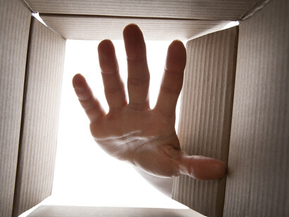 Una mano aperta e tesa si protende dall'alto verso l'interno di una scatola di cartone, creando un effetto di aspettativa