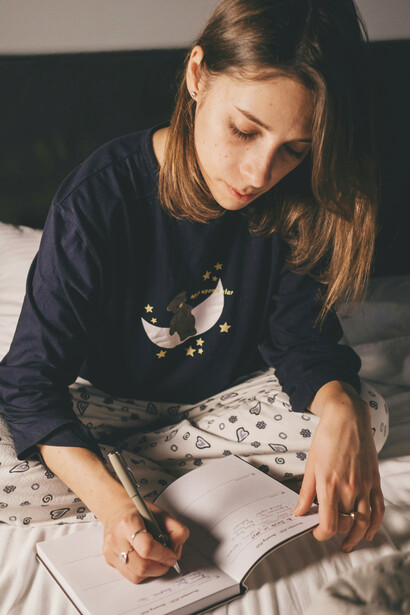 A young woman writing in a notebook while sitting comfortably on her bed, surrounded by a calm and intimate setting