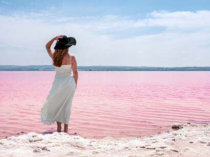 A woman wearing a white dress strolls along the edge of a breathtaking pink lake