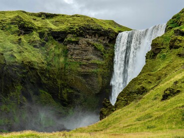 Vista da Skógafoss cercada pela paisagem verde e pelas formações rochosas da região, na cidade de Skógar, Islândia. Um lembrete de que a aventura islandesa vai muito além das atrações urbanas