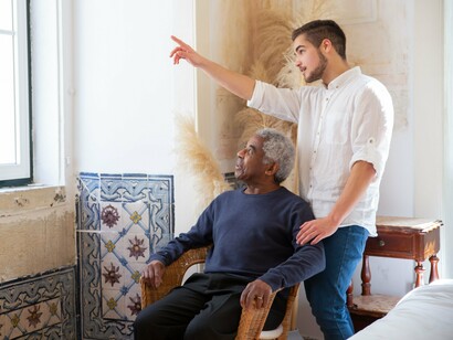 Em uma sala de estar, um homem mais jovem acompanha um homem idoso sentado em uma cadeira, diante de uma janela. A cena evoca a tranquilidade de uma presença compartilhada, sem necessidade de grandes gestos. Quando o vigor já não se expressa como antes, a vida continua a encontrar formas de presença, afeto e significado