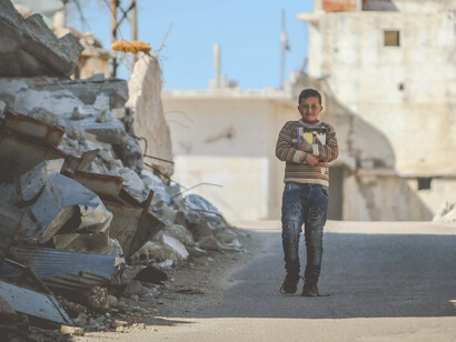 A young boy walks alone through a street lined with bombed-out houses, the devastation of war in Syria all around him