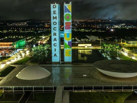 O Congresso Nacional do Brasil recebe a projeção da ação "Democracia Inabalada", que exibiu as mensagens "Democracia" e "Nos Une" nas torres do parlamento em memória de um ano dos ataques de 8 de janeiro de 2023. Em um cenário de incertezas políticas, o texto discute os desafios para preservar e renovar a democracia. Palácio do Congresso Nacional, Praça dos Três Poderes, Brasília - DF - Brasil