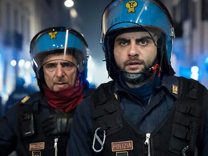 The riot police gather in silence, their gear reflecting the harshness of their duty, but their eyes betray the human complexities that fuel their actions on the streets