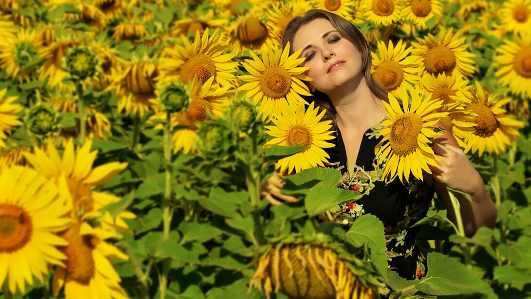 A woman in a black and white floral shirt stands in a sunflower field under the bright daylight, radiating inner peace and a positive mindset