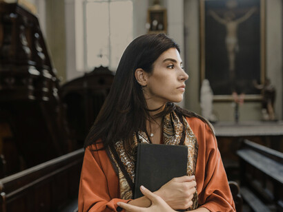 Woman in an orange long-sleeve shirt praying in church with a black Bible in her hands