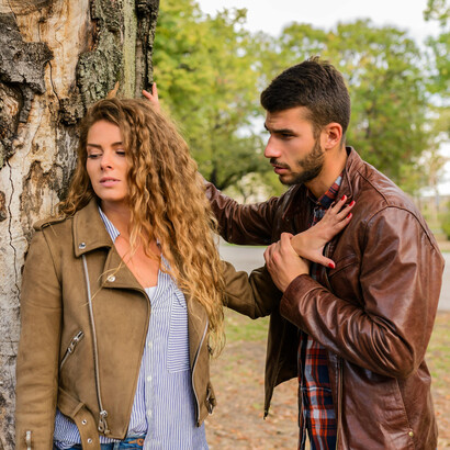 A man and woman wearing brown jackets stand near a tree, their silence and distance reflecting the emotional disconnect and loneliness between them