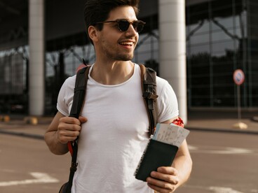 A young male traveler wearing a white T-shirt and sunglasses, smiling near an airport