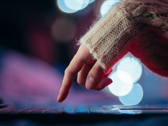 Uma mão pressiona uma tecla de um computador portátil, cercada por pontos de luz desfocados. Um gesto mínimo, quase imperceptível, pode representar o poder de acionar sistemas inteiros — uma metáfora para os riscos de um mundo financeiro cada vez mais dependente de comandos digitais