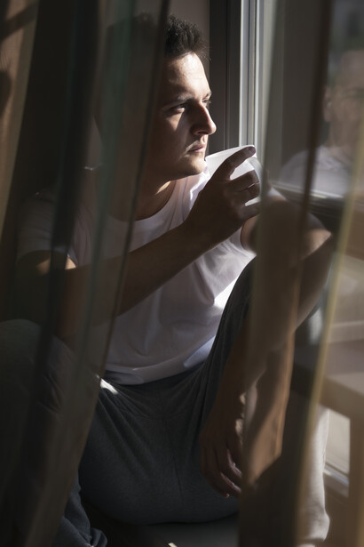 A man sits by a window, soft light filtering through the curtains as shadows fall across his face while he reflects quietly