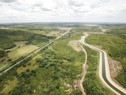 Um canal do Projeto de Integração do Rio São Francisco atravessa a paisagem do semiárido, cercado pela vegetação e por áreas de ocupação humana. A obra representa a capacidade da engenharia de modificar a relação entre a água e o território