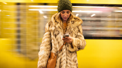A young woman stands at a train station while staring at her phone, reflecting social isolation and digital disconnection in modern urban society