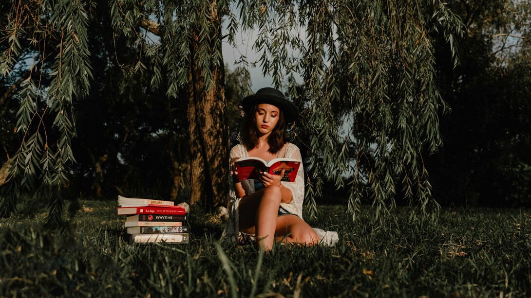 Jovem lê um livro sob a sombra de uma árvore, cercada por outros livros. A imagem remete ao início de uma busca interior que une contemplação, conhecimento e silêncio