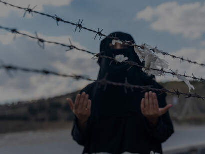 A refugee woman stands behind a fence, her posture tense, as she looks toward an uncertain future