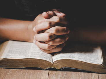 A close-up of folded hands resting on a Holy Bible inside a softly lit church, reflecting Christian faith and devotion